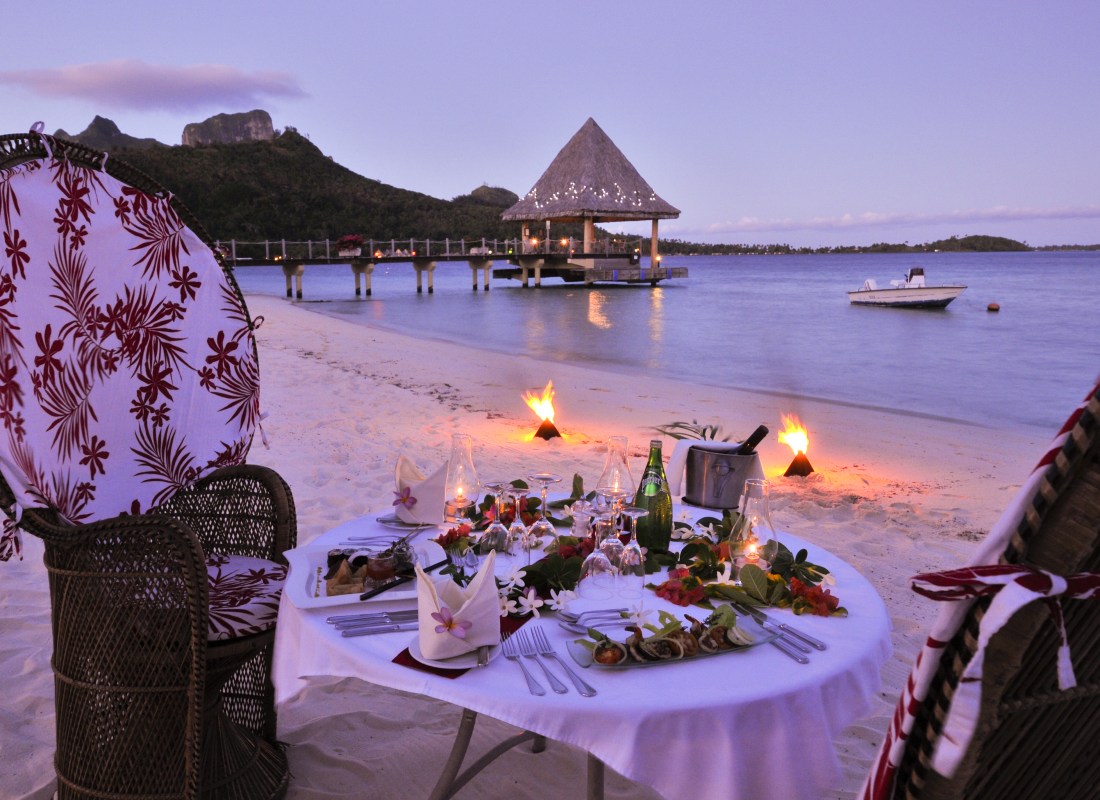 A table is laid out with food and champagne and it sits on the beach at night with fire. 